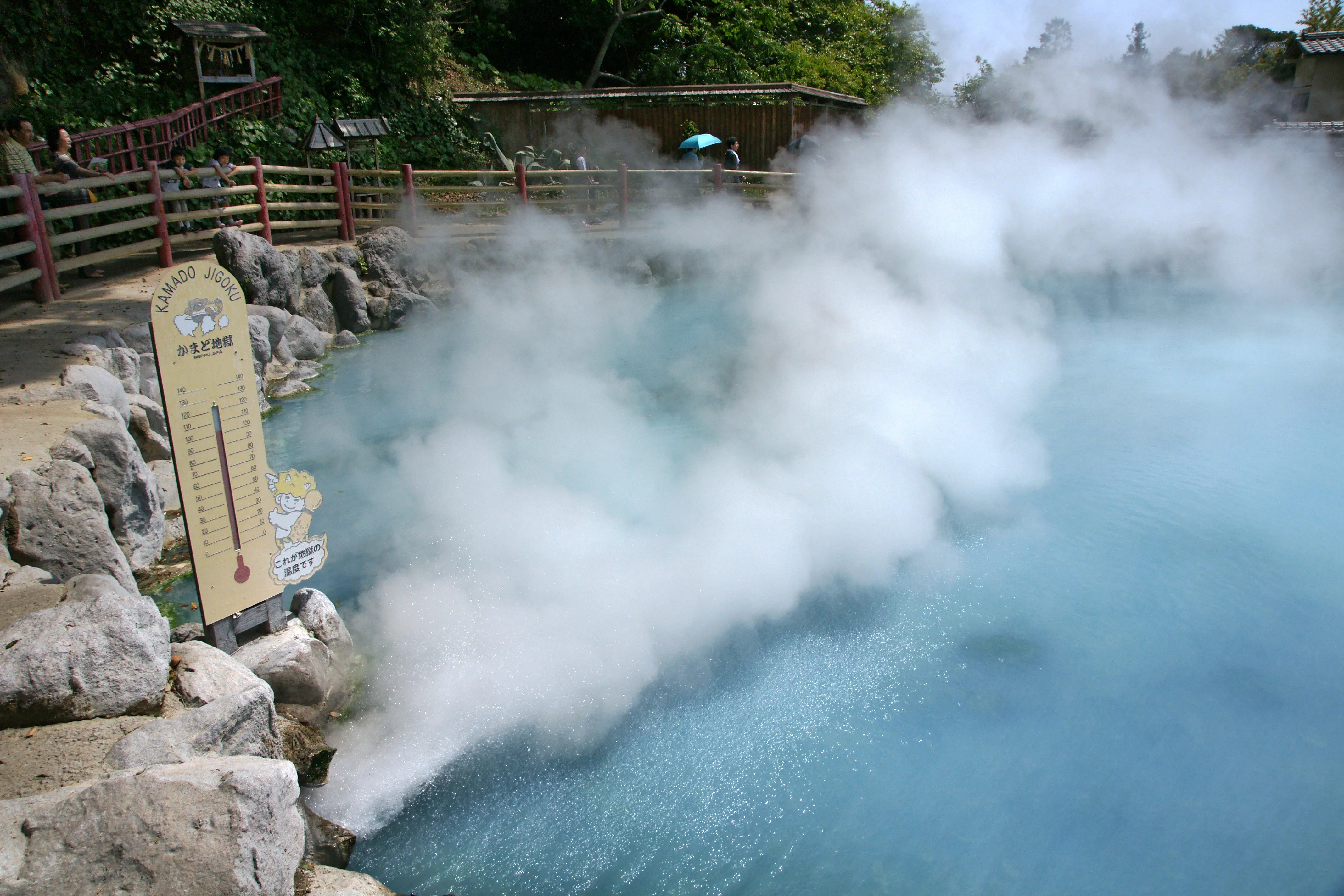 Steam pouring across the pale blue pool of Kamado Jigoku, the Cooking Pot Hell, with its painted thermometer signboard on the rocks and visitors at the railing
